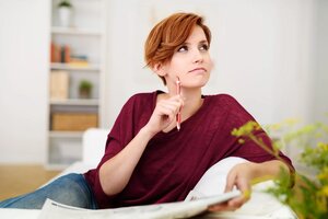 Thoughtful Attractive Young Woman Answering Crossword Puzzle Game on Newspaper at the Living Room Couch